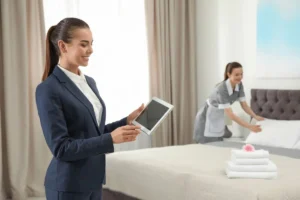 Hotel cleaner making a bed as the housekeeping supervisor reviews tasks on a tablet.