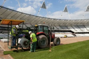 Two grounds maintenance workers using a tractor to lay grass on a football field.