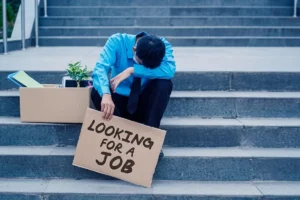 Unemployed man holding a "Looking for Job" sign while sitting with his head down.